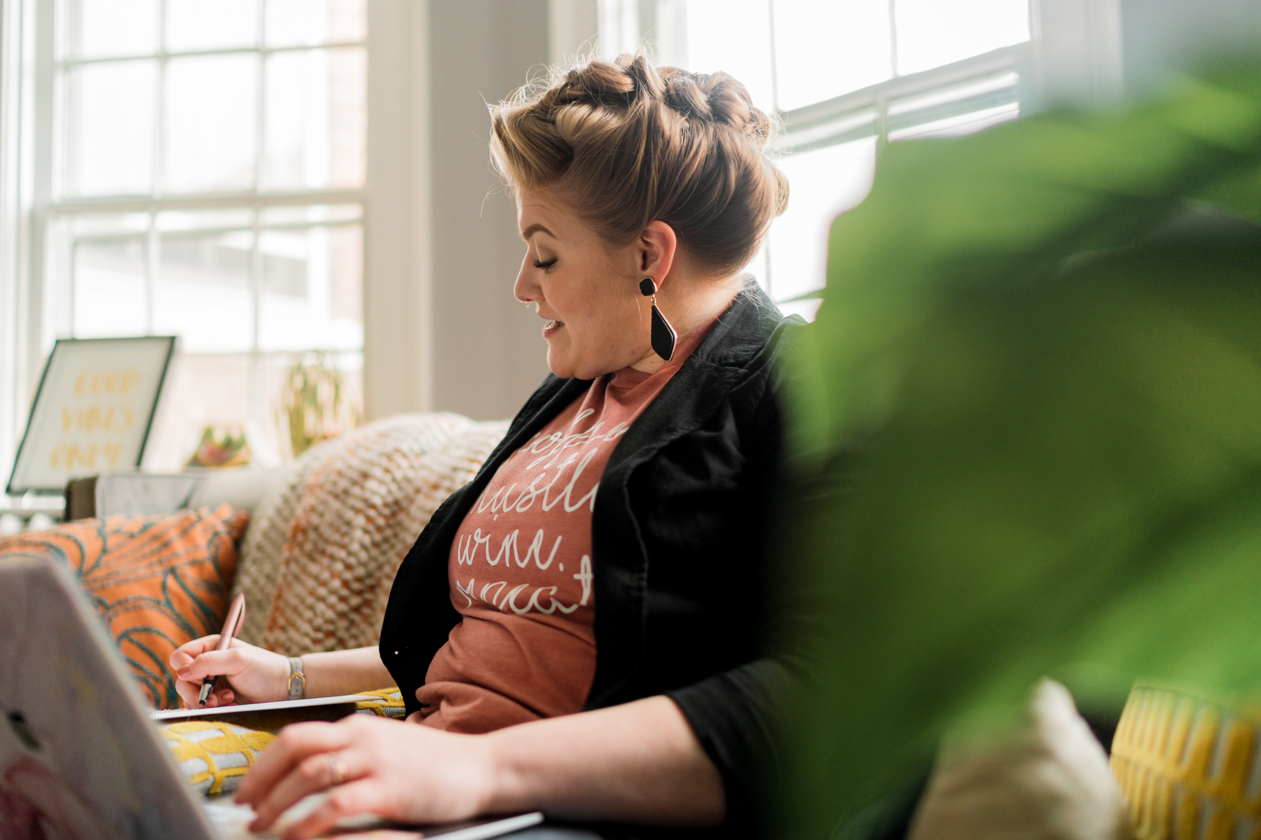 Side view of of Becca Mai, a wedding business coach, sitting on a couch working on her laptop in her lap while taking notes in a spiral-bound notebook propped on a pillow