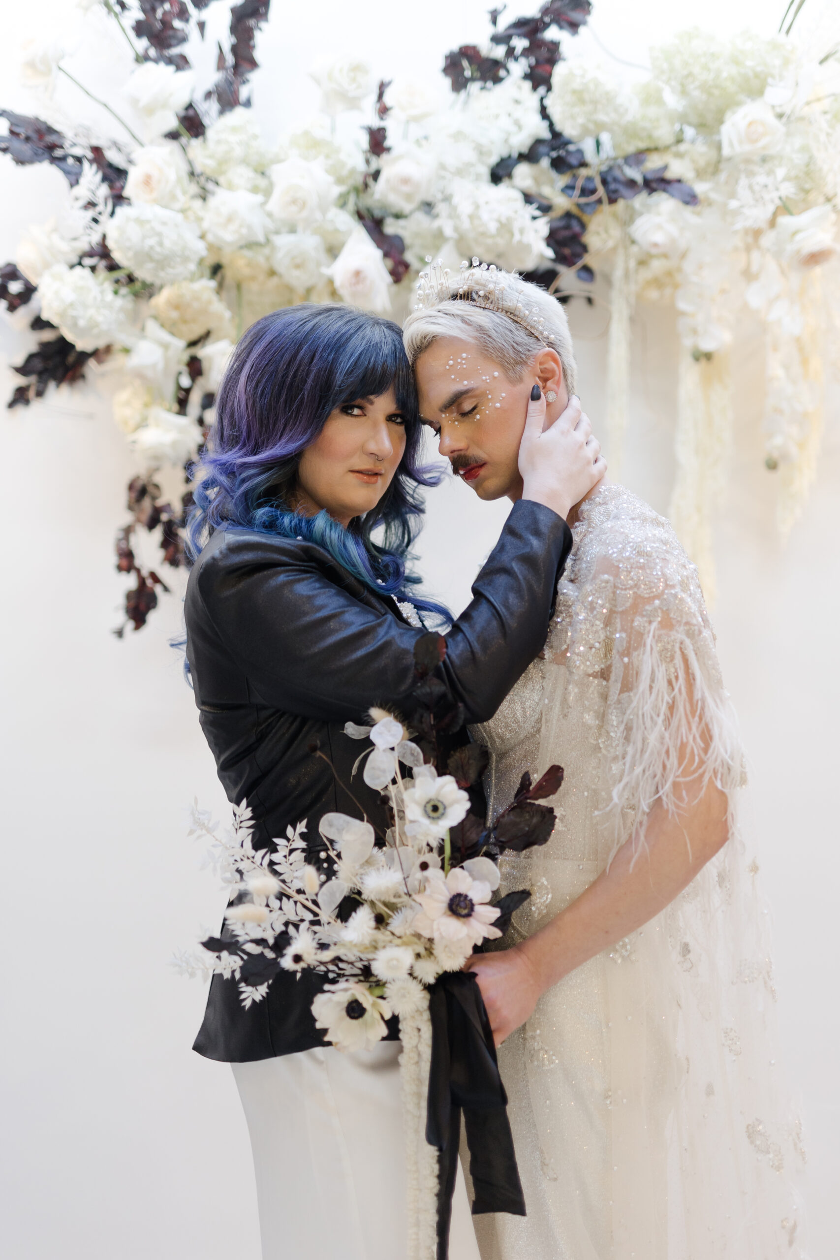 nonbinary marriers in front of ceremony altar with one marrier in a black blaser with pearls on the lapel and other marrier in an ivory bejeweled jumpsuit