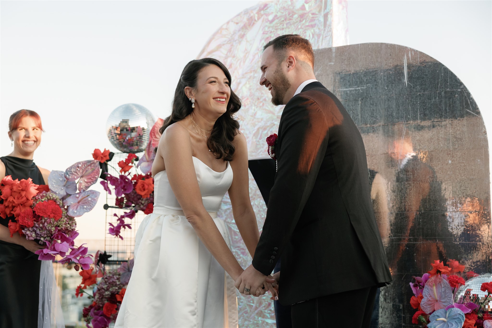 hetero-cis couple holding hands in front of iridescent and silver sequin backdrop surrounded by vibrant colorful pink, red, and purple flowers and disco balls