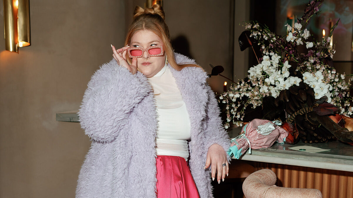Becca Mai a wedding business coach leaning on a bar at Ammora in Baltimore posing for the camera in an Anthropologie lavender fuzzy coat with pink silk skirt and white sheer top looking off in the distance while holding her pink and pearl sunglasses on the bridge of her nose