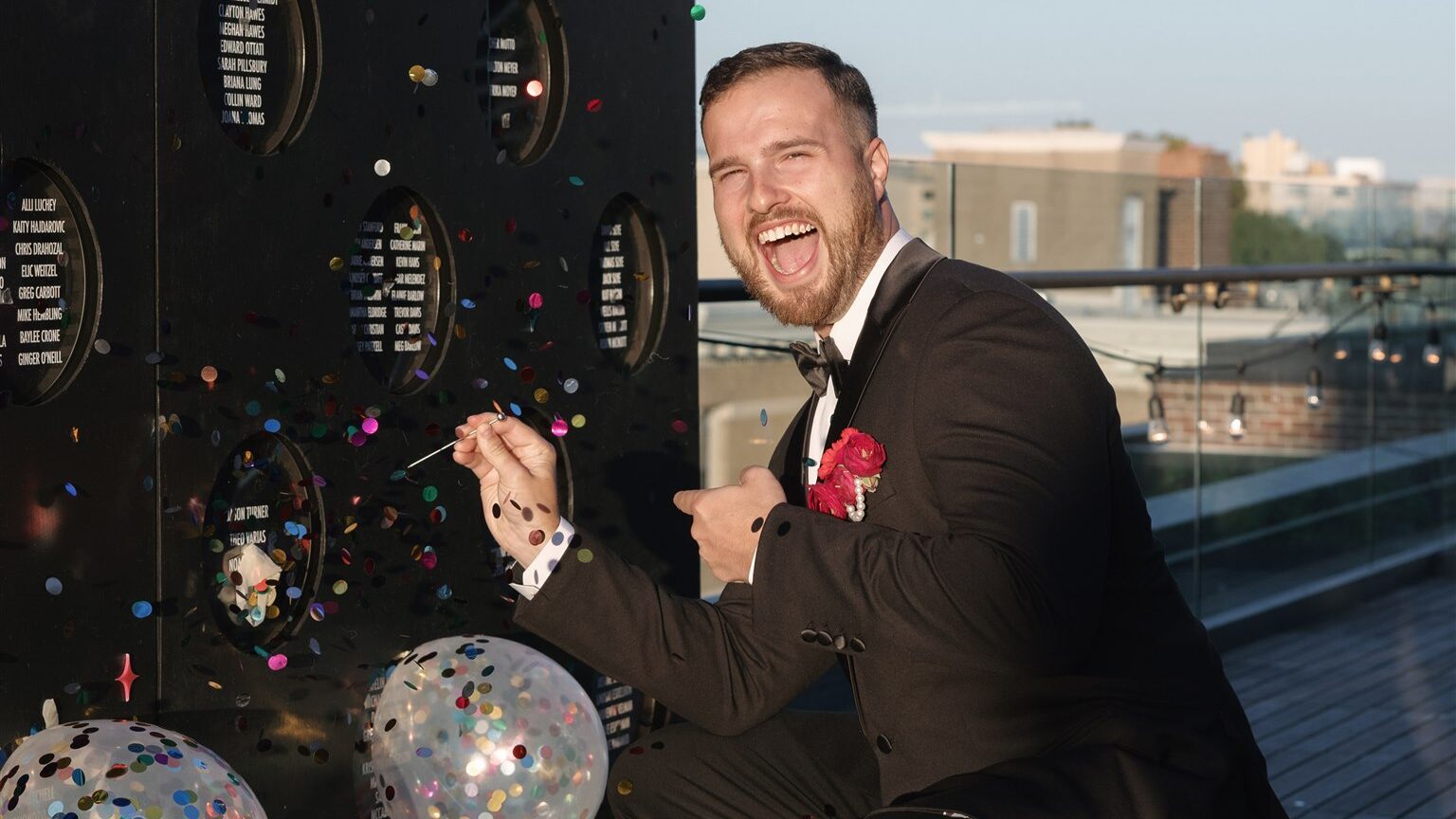 Groom squatting down, smiling, and laughing at the camera while he pops a confetti balloon on his escort wall on the terrace at the Line Hotel DC