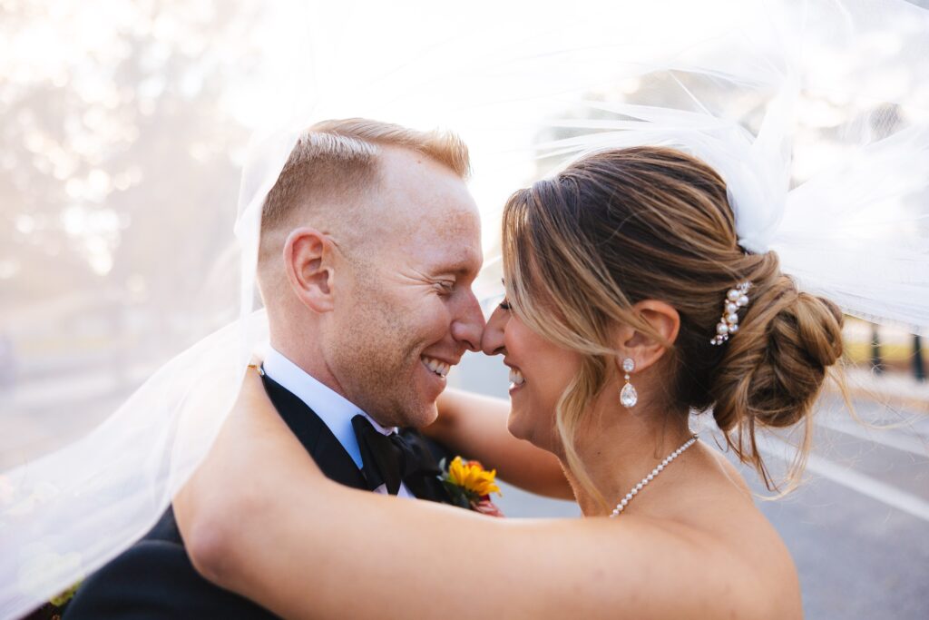 bride holding groom around the neck and both are touching noses, eyes closed, and smiling with brides veil over both of their heads on a street in Capitol Hill DC
